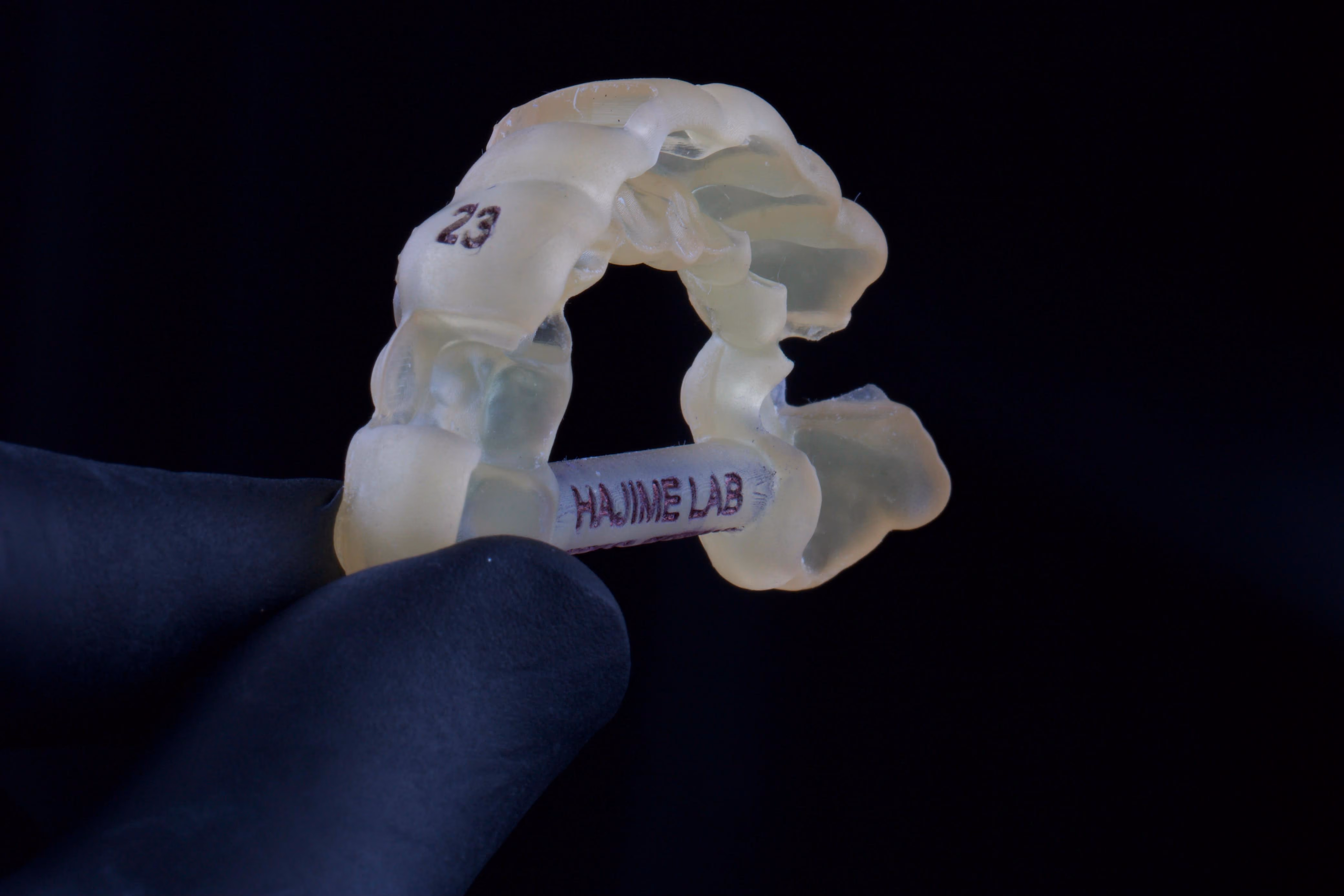 Guia cirúrgico impresso em resina translúcida, segurado por uma mão com luva preta, com a marcação do dente 23 e a gravação Hajime Lab
