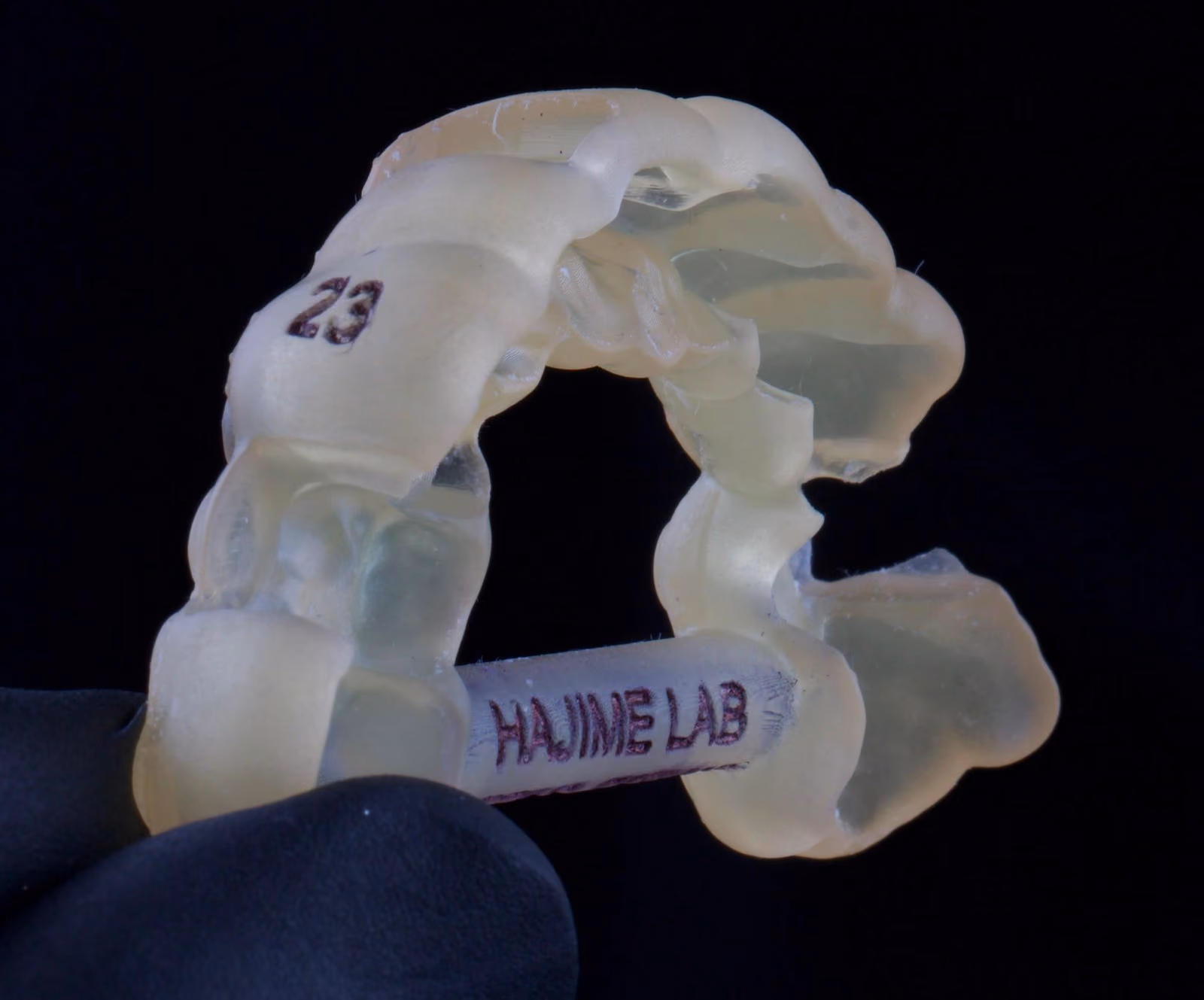 Guia cirúrgica impressa em resina translúcida, segurada por uma mão com luva preta, com a marcação do dente e a gravação Hajime Lab.